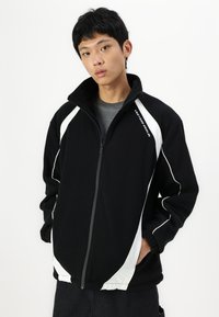Giacca in fleece nera con zip, dettagli e logo bianchi, con colletto alto e vestibilità rilassata. Materiale testurizzato, maniche lunghe.