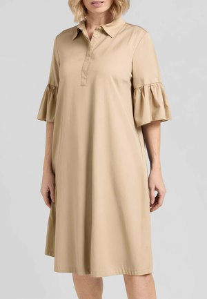 Donna che indossa un vestito beige al ginocchio con maniche a volant al gomito e colletto abbottonato, in piedi davanti a uno sfondo semplice.