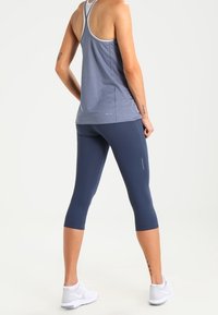 Tmavě modré tříčtvrteční legíny a světle modrý top s průstřihy na zádech, oba přiléhavé, s jemnými logy a hladkou, texturovanou látkou. Bílé sportovní boty.