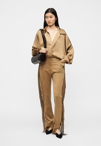 Μπεζ σατέν πουκάμισο με φαρδιά μανίκια, σετ με μπεζ παντελόνι με πλαϊνές φούντες. Το μοντέλο κρατά μια στρογγυλή μαύρη τσάντα και φορά μαύρα τακούνια.