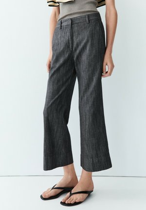 Personne portant un pantalon large gris foncé raccourci, un haut sans manches beige et des sandales tongs noires, debout devant un fond blanc.