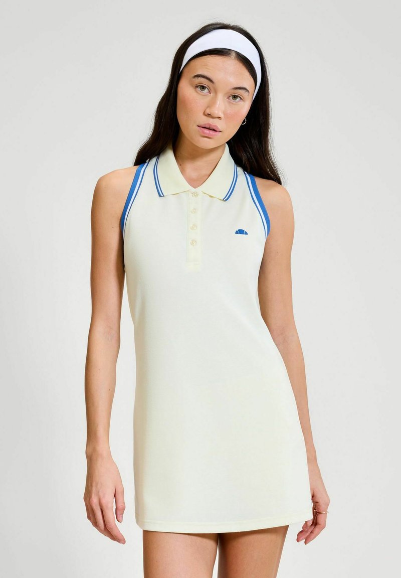 Donna con capelli lunghi e scuri che indossa un abito polo crema senza maniche con bordi blu e una fascia bianca per capelli, in piedi davanti a uno sfondo semplice.