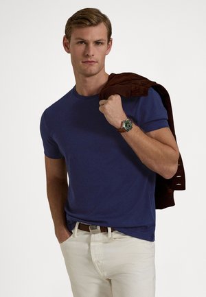 Μπλε ναυτικό βαμβακερό t-shirt με στρογγυλή λαιμόκοψη, σε συνδυασμό με λευκό παντελόνι. Καφέ μπουφάν ριγμένο στον ώμο, ασημένιο ρολόι με πράσινο καντράν στον καρπό.