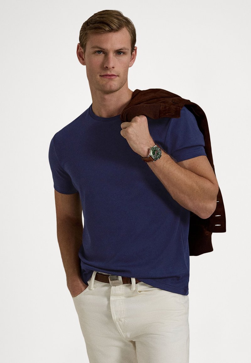 Maglietta blu navy con scollo tondo in cotone, abbinata a pantaloni bianchi. Giacca marrone appoggiata sulla spalla, orologio d'argento con quadrante verde al polso.