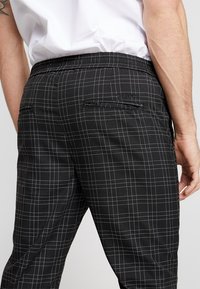 Svart rutiga byxor med vitt rutmönster, med en elastisk midja och en enda bakficka. Slät tygtextur, skräddarsydd design.