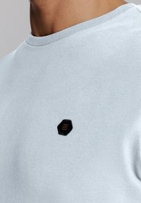Helles blaues Sweatshirt mit Rundhalsausschnitt, weicher Textur und einem hexagonalen schwarzen Patch mit goldenen Streifen auf der Brust.