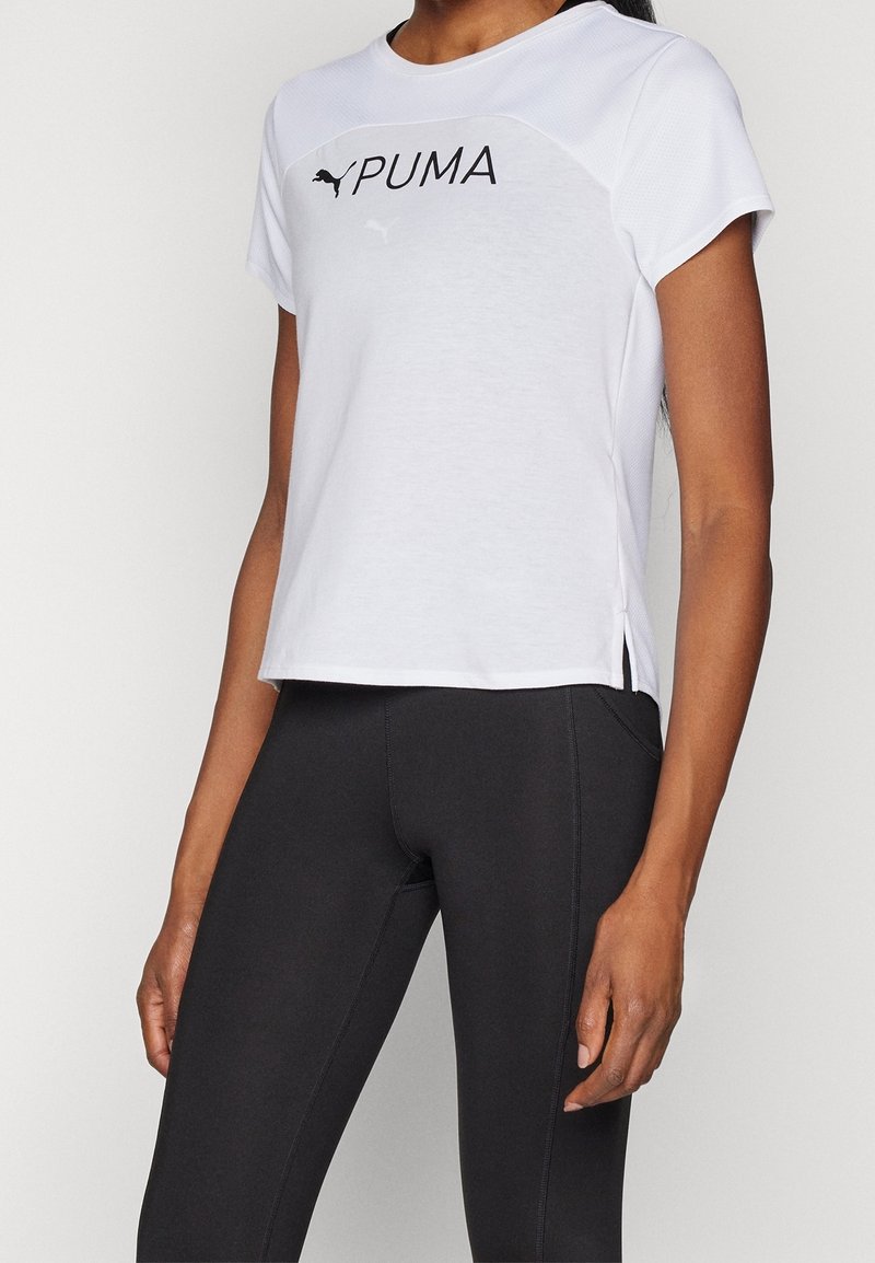 Camisa deportiva blanca de manga corta con el logo negro de "PUMA", escote redondo y cortes laterales. Combinada con leggings negros, que destacan un diseño ajustado.