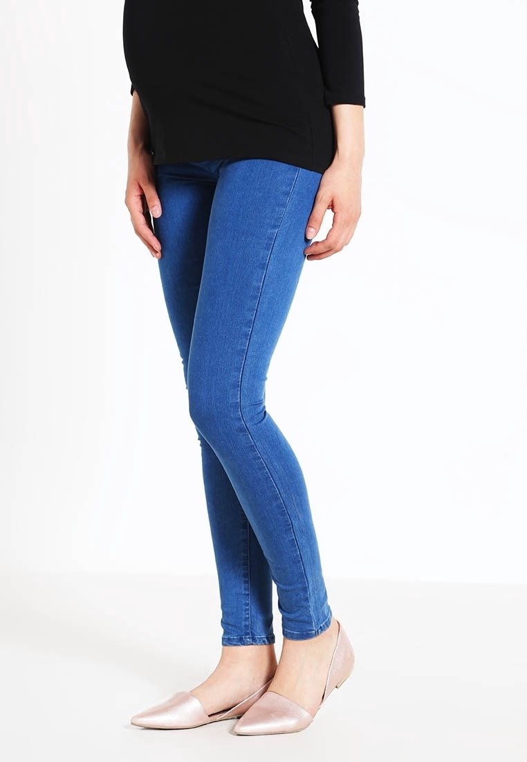 Femme portant un jean skinny bleu, un haut noir et des ballerines pointues rose clair, debout sur un fond blanc.