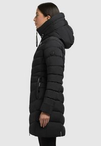 Un cappotto imbottito nero con colletto alto e cappuccio, caratterizzato da un design trapuntato, tasche laterali con zip e una patch con logo sulla manica.