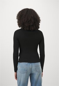 Persona con cabello rizado de pie de espaldas, vistiendo una camiseta de manga larga negra con relieve y jeans de mezclilla azul claro.