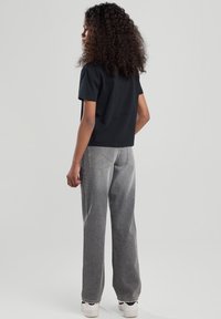 Camiseta corta negra de manga corta combinada con jeans grises de corte holgado. El modelo lleva zapatillas blancas y se puede ver su cabello rizado por detrás.
