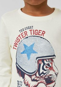 Cremfarbendes Langarmshirt mit einem Grafikdesign eines Tigers, der einen silbernen Helm mit einem blauen Stern trägt. Darauf steht der Text "DER GROßE TWISTER TIGER."