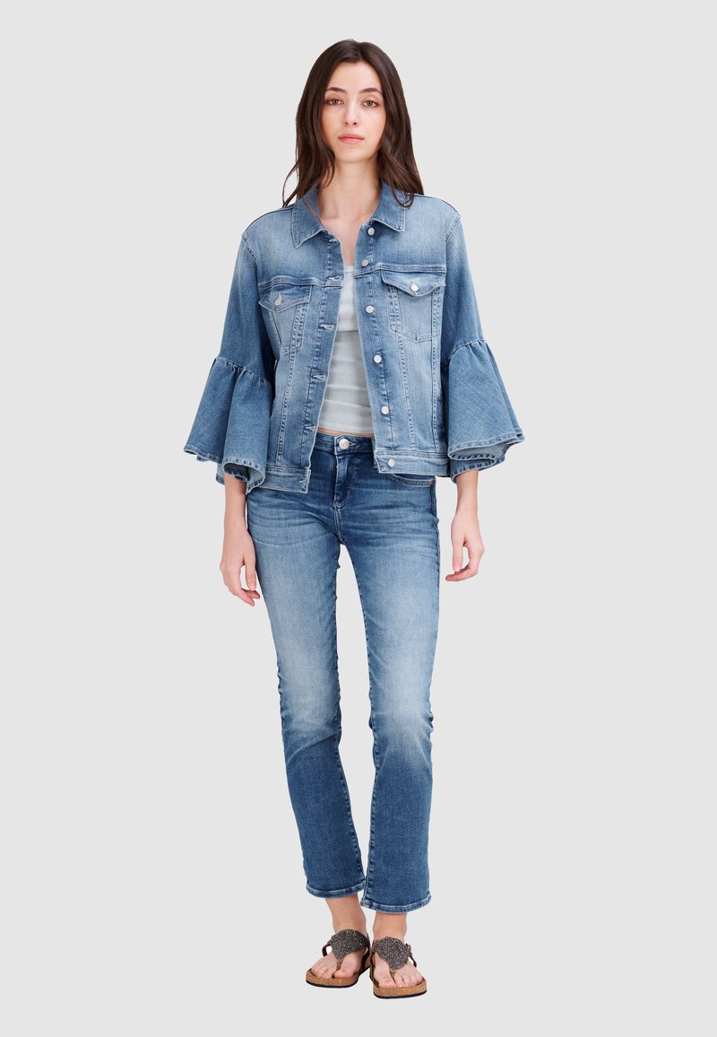 Jeansjacke mit ausgestellt Ärmeln, hellblauer Farbe, Fronttaschen und silbernen Knöpfen. Kombiniert mit enganliegenden Jeans und dekorativen Sandalen.