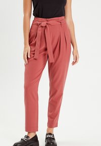 Pantalons taille haute de couleur rosé avec une ceinture nouée à l'avant, comprenant des plis et un design de jambe fuselée. Portés avec des mocassins noirs.