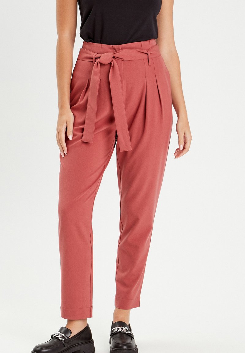 Pantalons taille haute de couleur rosé avec une ceinture nouée à l'avant, comprenant des plis et un design de jambe fuselée. Portés avec des mocassins noirs.