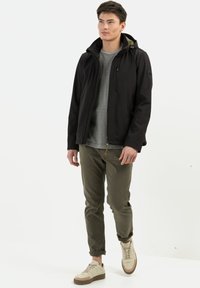 Zwarte softshell jas voor heren met een capuchon, textuurstof en een ritszak, gecombineerd met een grijs shirt en olijfgroene broek, crème sneakers.