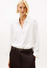 Kvinde med kort, blond hår iført en hvid skjorte med knapper, stoppet ind i sorte bukser med et sort bælte, står foran en ensfarvet baggrund.