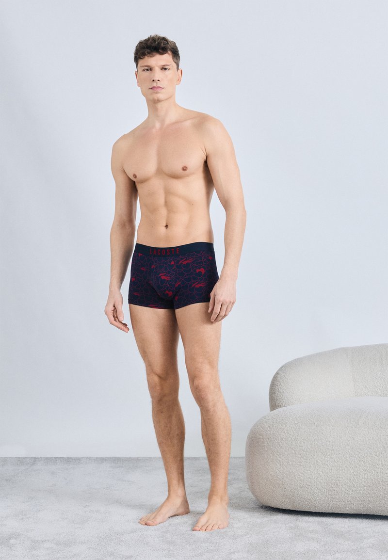 Άνδρας χωρίς πουκάμισο στέκεται ξυπόλητος πάνω σε χαλί, φορώντας ναυτικό μπλε πυτζάμες με κόκκινο φλοράλ σχέδιο και ζώνη μέσης "Lacoste", δίπλα σε καμπυλωτό μπεζ καναπέ.