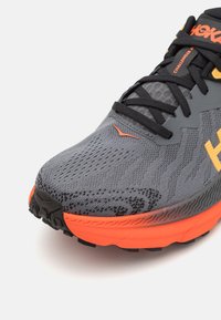 Šedá atletická bota s oranžovými akcenty, vybavená texturovaným pleteným svrškem, podporujícími překryvy a odolnou černou podešví s protiskluzovým vzorem.