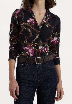 Kvinde iført sort langærmet skjorte med lilla blomster- og guldkædemønster, stukket ned i mørkeblå jeans med brunt bælte.