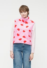 Γούνινο γιλέκο σε ροζ χρώμα με κόκκινο μοτίβο καρδιάς, διαθέτοντας ψηλό γιακά και μπροστινές τσέπες. Φορεμένο πάνω από ένα ανοιχτό πουλόβερ και συνδυασμένο με τζιν παντελόνι.