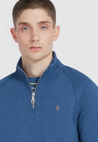 Blue zip-up pullover made of soft fabric, featuring a stand collar, a silver zipper, and an orange logo on the chest.