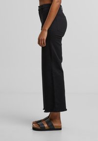 Person iført sorte jeans med højt talje og lige ben med rå kant og sorte platform-sandaler, stående sidelæns mod en ensfarvet grå baggrund.