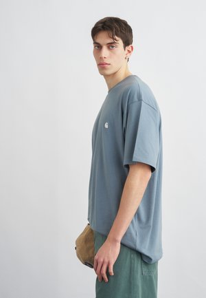 Joven con cabello corto y oscuro, vistiendo una camiseta oversize azul grisácea y pantalones verdes, sosteniendo una gorra beige sobre un fondo liso.