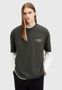 Camiseta de algodón verde oscuro con cuello redondo, mangas cortas y logotipo blanco en el pecho, superpuesta sobre una camisa blanca de manga larga.