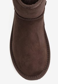 Brązowy zamszowy botek za kostkę z teksturowaną powierzchnią, o zaokrąglonym kształcie i wzmocnioną tkaniną na górze. Wzmocnione przeszycia wzdłuż podeszwy.