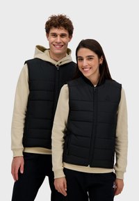 Czarne kamizelki puchowe z zamkiem z przodu, z pikowanymi panelami i bocznymi kieszeniami. Noszone na kremowych bluzach z kapturem, co podkreśla swobodny styl.