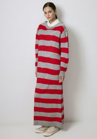 Rochie supradimensionată cu dungi roșii și gri, având glugă, mâneci lungi, manșete cu nervuri și lungime maxi. Purtați cu adidași bej.