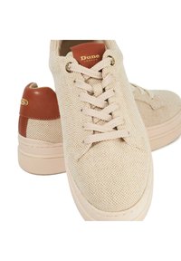 Baskets en toile beige avec une surface texturée, présentant un accent de talon en cuir marron et des lacets blancs. Semelles en caoutchouc avec une adhérence à motifs.