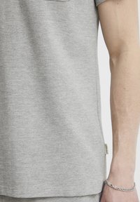 Graues, strukturiertes T-Shirt mit kurzen Ärmeln, das eine Taschen und ein kleines Etikett an der Seite hat. Glattes Gewebe mit einem dezenten gerippten Muster.