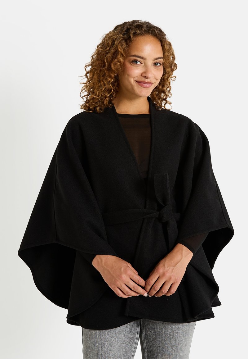 Wrap de mezcla de lana negra con un diseño de capa estructurada, que presenta mangas anchas y un detalle de cinturón anudado, combinado con pantalones grises texturizados.