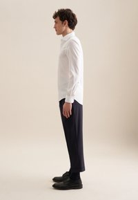 Camicia bianca con bottoni, pantaloni navy a vita alta, scarpe nere. La camicia ha un colletto classico e maniche lunghe con dettagli a bottoni. Tessuto dalla superficie liscia.