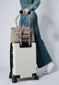 Valigia rigida bianca a coste con accenti neri e ruote, sormontata da una borsa in pelle beige con tasca zip e tracolla stampata.