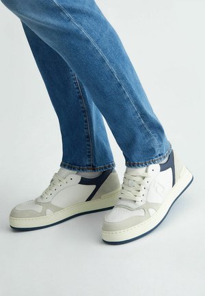 Sneakers bianchi e grigi con accenti blu navy, texture lisce e in suede, punta rotonda, suole piatte e cuciture a contrasto, abbinati a jeans in denim.