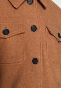 Brun uldfrakke med sorte knapper, to klapbrystlommer og en spidse krave, vist i tæt detalje.