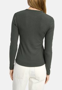 Ciemnozielona, prążkowana bluzka z długim rękawem, okrągłym dekoltem, dopasowanym krojem i gładką fakturą, noszona z jasnymi dżinsami.