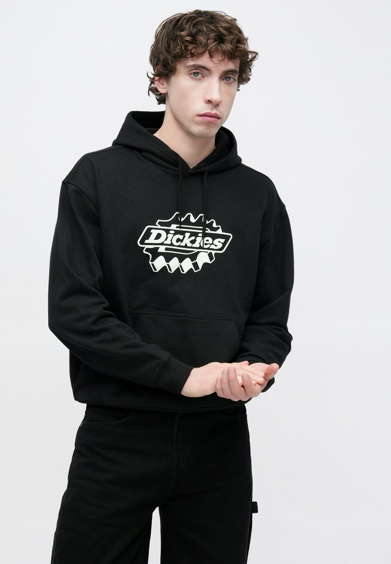 Sudadera negra con capucha, bolsillo delantero tipo canguro y logo blanco de "Dickies" con bordes serrados. La tela parece suave y con textura. Combinada con pantalones negros.