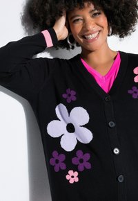 Schwarzer Cardigan mit großen lila und rosa Blumenmustern, V-Ausschnitt und drei schwarzen Knöpfen. Mit kontrastierenden rosa Manschettenakzenten.