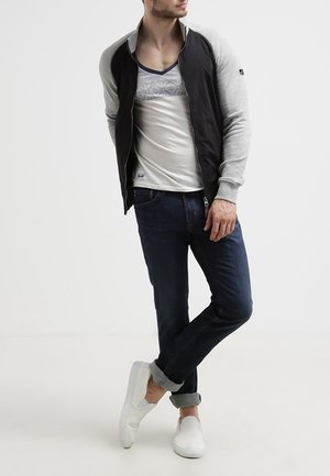 Czarna kurtka bomberka na zamek z szarymi rękawami, noszona na białej koszuli z geometrycznym wzorem. Zestawiona z ciemnoniebieskimi dżinsami i białymi butami slip-on.