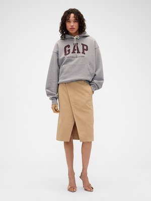 Mujer con sudadera con capucha gris GAP Victoria Beckham, falda cruzada beige hasta la rodilla y zapatos de tacón nude con tiras, posando sobre un fondo blanco liso.