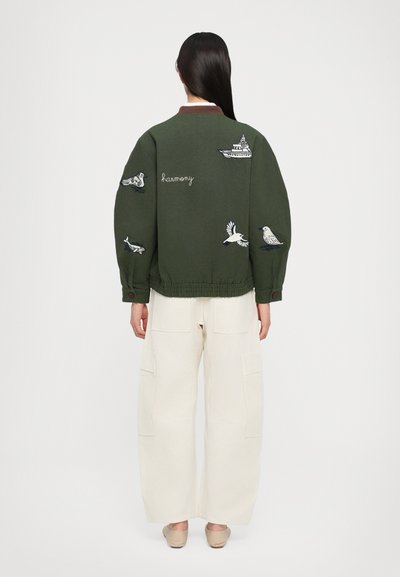 Chaqueta bomber verde con bordados de pájaros y un diseño de barco. Presenta un dobladillo ajustado y un cuello de canalé, combinada con pantalones de pierna ancha beige.