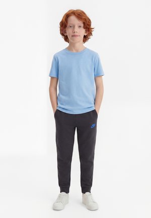 Dieťa s červenými vlasmi stojí s rukami v vreckách, oblečené v svetlomodrom tričku, čiernych joggeroch Nike a bielych teniskách na jednofarebnom pozadí.