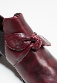 Chaussure en cuir bourgogne avec un détail de nœud et une finition texturée. Caractérisée par un petit talon et un panneau élastique pour un enfilage facile.