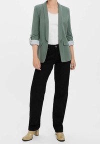 Ljusgrön kavaj med en elegant design, uppvikta ärmar med randiga detaljer, kombinerad med svarta byxor och beige klackskor.