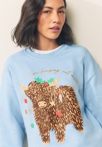 Jasnoniebieska bluza z nadrukiem fakturowanej, brązowej krowy wysokogórskiej ozdobionej świątecznymi ozdobami i zielonymi akcentami ostrokrzewu. Krótkie rękawy, luźny krój.