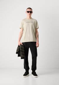 Lichtbeige t-shirt met Calvin Klein-logo, zwarte cargo-broeken en zwarte sneakers, met een gestreepte donkere trui in de hand. Zwarte zonnebril.